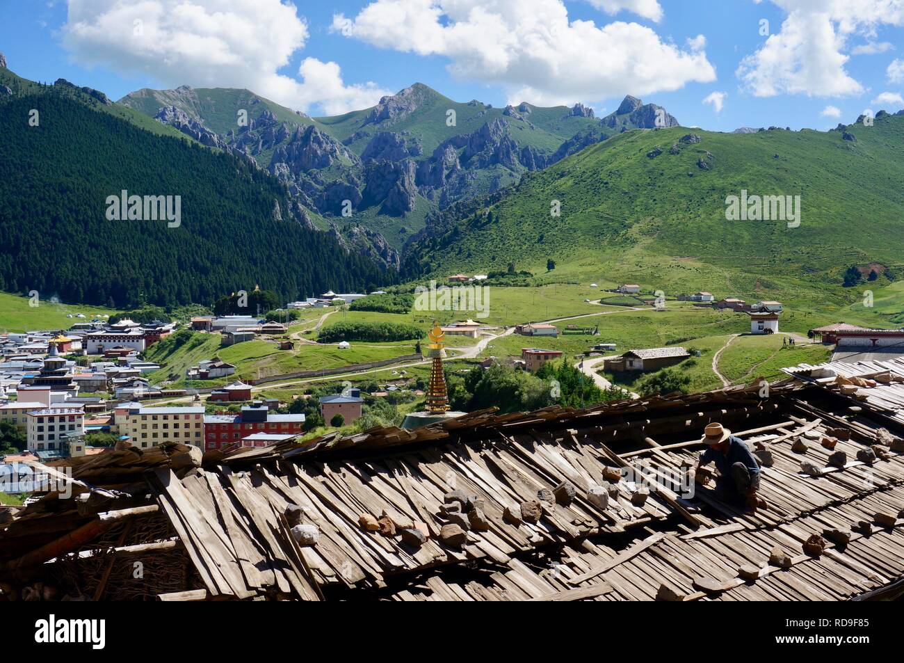 Ein Mann Reparaturen einen alten Tempel auf der Dachterrasse mit Blick auf eine moderne Stadt- und Berglandschaft hinter ihm. Langmusi Kloster, China Stockfoto