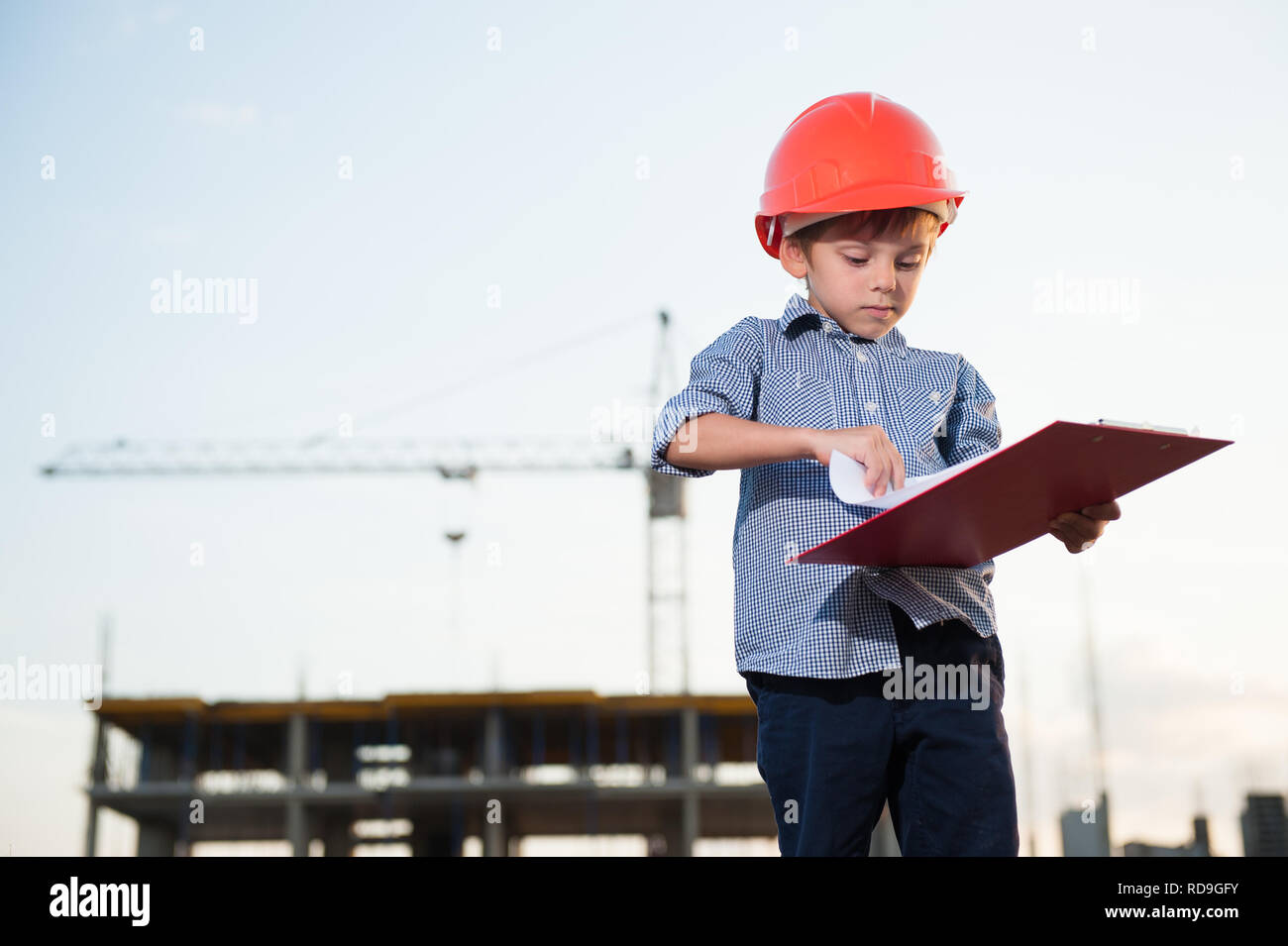 Cute little boy in orange Helm mit Datei mit Projekt Blätter auf der Baustelle mit Kran Stockfoto