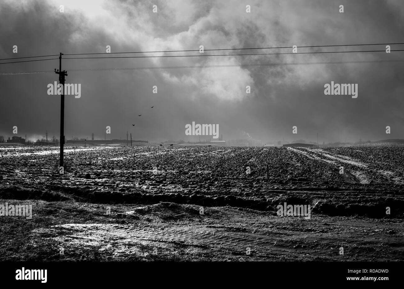 Blick auf die gefrorene gepflügten Feldes, die Strahlen der Sonne durch die dramatische Wolken, industrielle Gebäude brechen in der Ferne zu sehen ist. Schwarz und whit Stockfoto