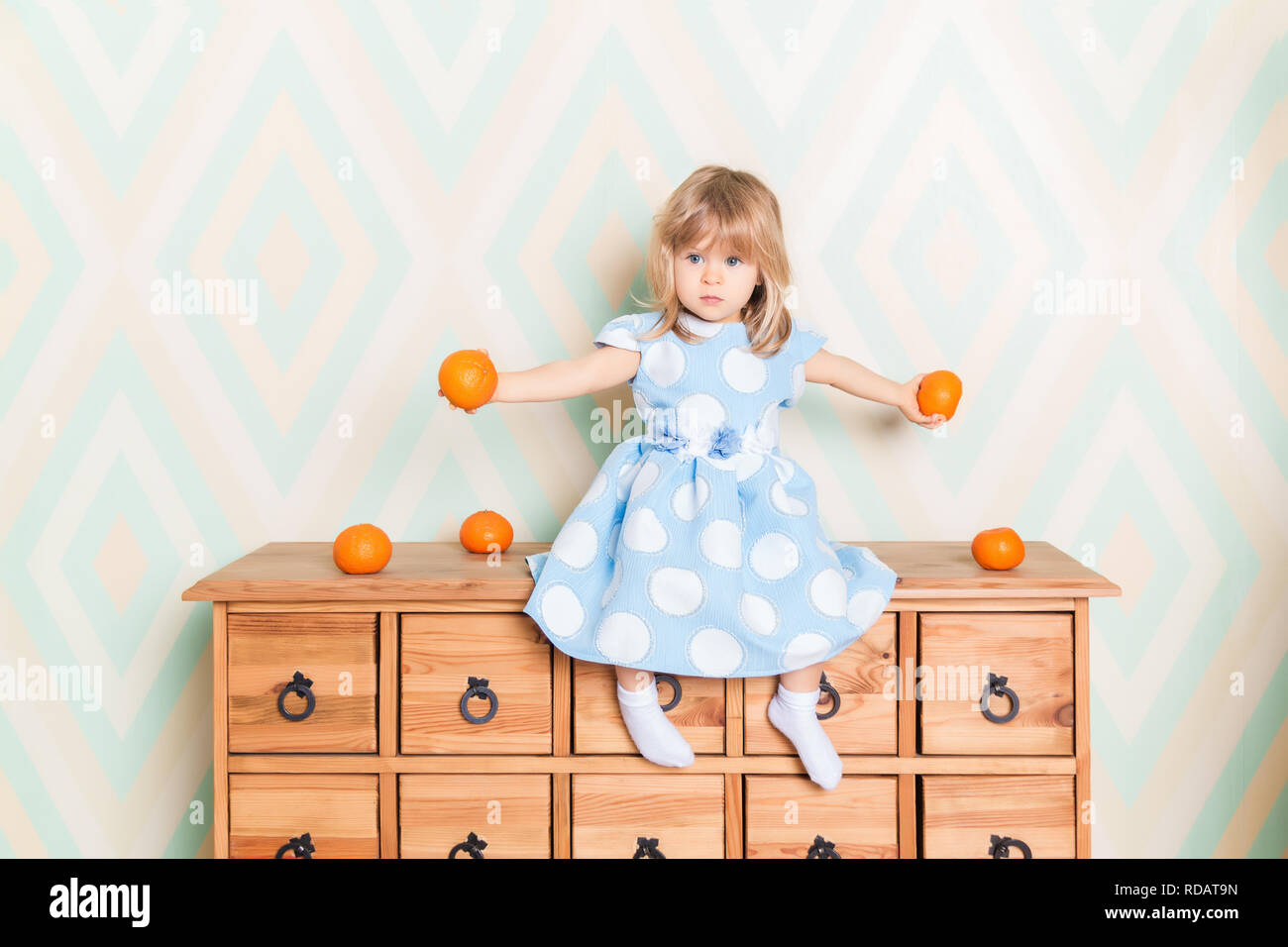 Kleinkind Kind Baby Mädchen im hellblauen Kleid Ernst sitzen auf die  hölzerne Kommode und halten frische Orangen Mandarinen in ihre Hände auf  Raute Hintergrund. Kinder- Emotionen Gefühle Stockfotografie - Alamy
