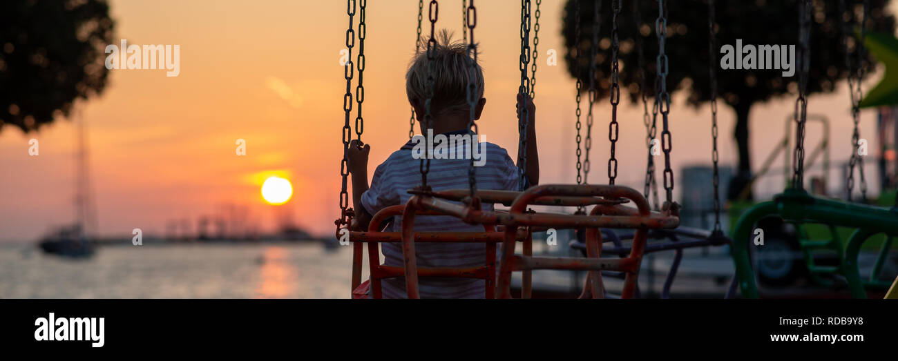 Ansicht von hinten von einem Kleinkind junge Reiten auf einem Karussell am Meer gegen einen schönen rot glühenden Sonnenuntergang. Stockfoto