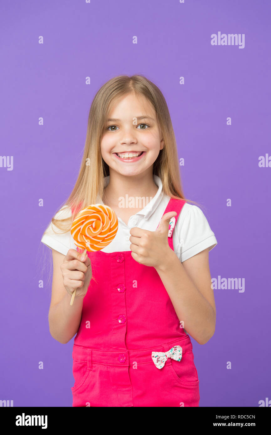 Mädchen mit bunten Lutscher. Süße Kindheit Konzept. Kind mit langen Haaren gerne Süßigkeiten. Mädchen mit lächelnden Gesicht hält Giant candy in der Hand, violett unterlegt. Kind in rosa Overalls, Daumen hoch. Stockfoto