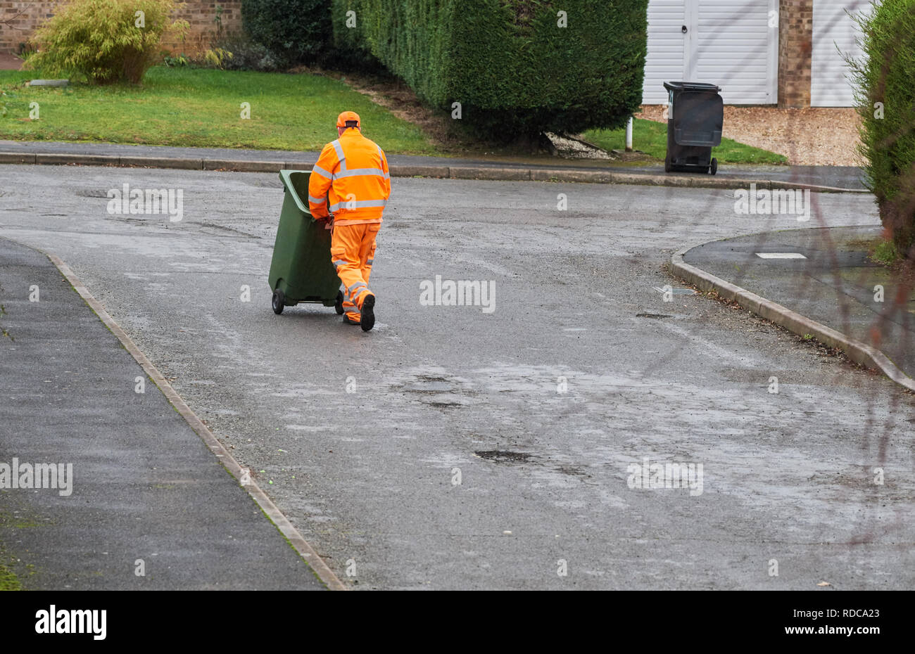 Ein Fach Mann in einem orange fluoreszierende Jacke und Hose bekleidet schiebt eine grüne recyceln Mülltonne mit wiederverwertbaren Abfalls auf einer Straße in einem nassen Winter Stockfoto