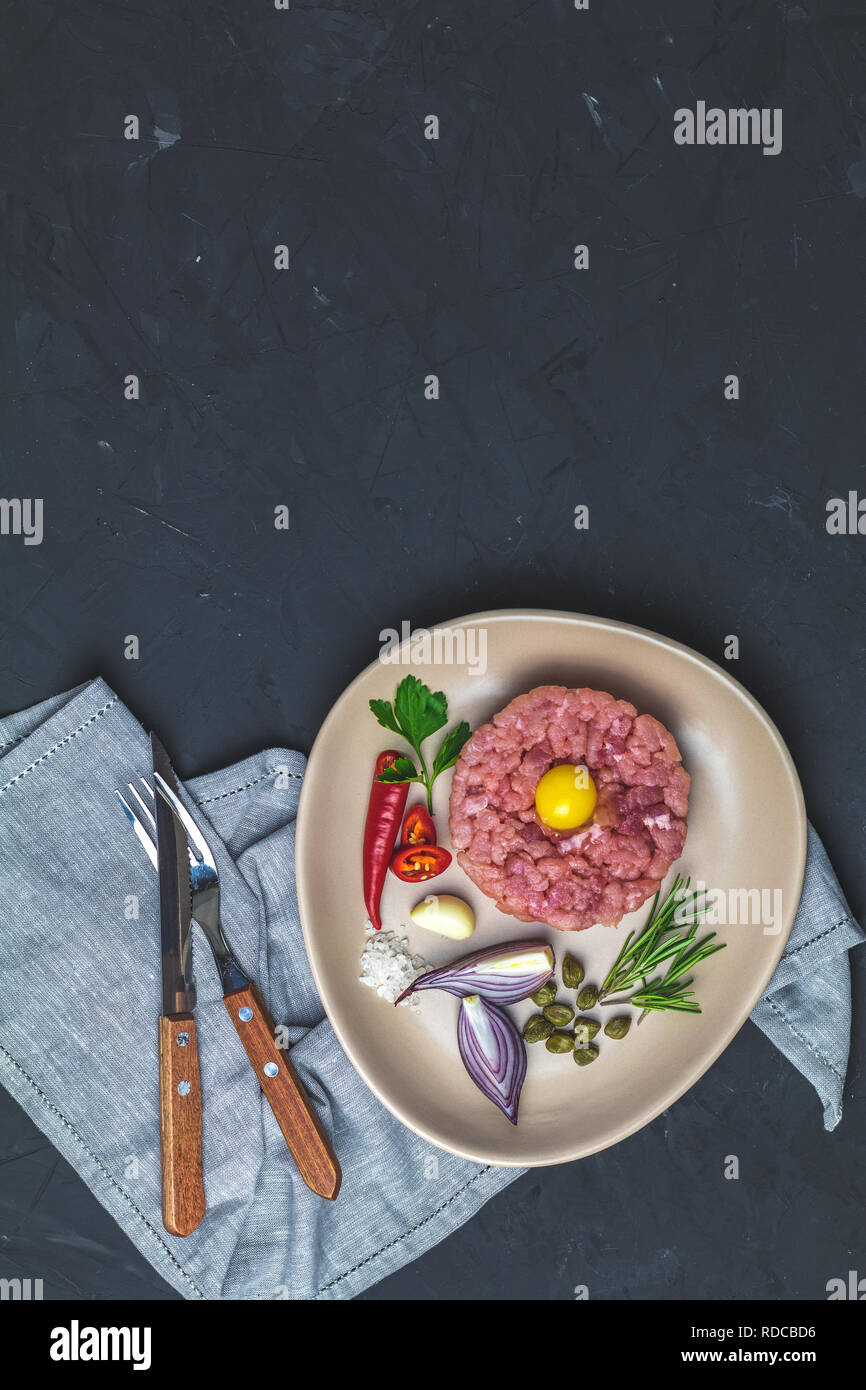Lecker Steak Tartar mit Eigelb und Zutaten auf Keramik Teller, Besteck Messer, Gabel auf schwarzen Stein Beton texturierte Oberfläche Hintergrund. Co Stockfoto