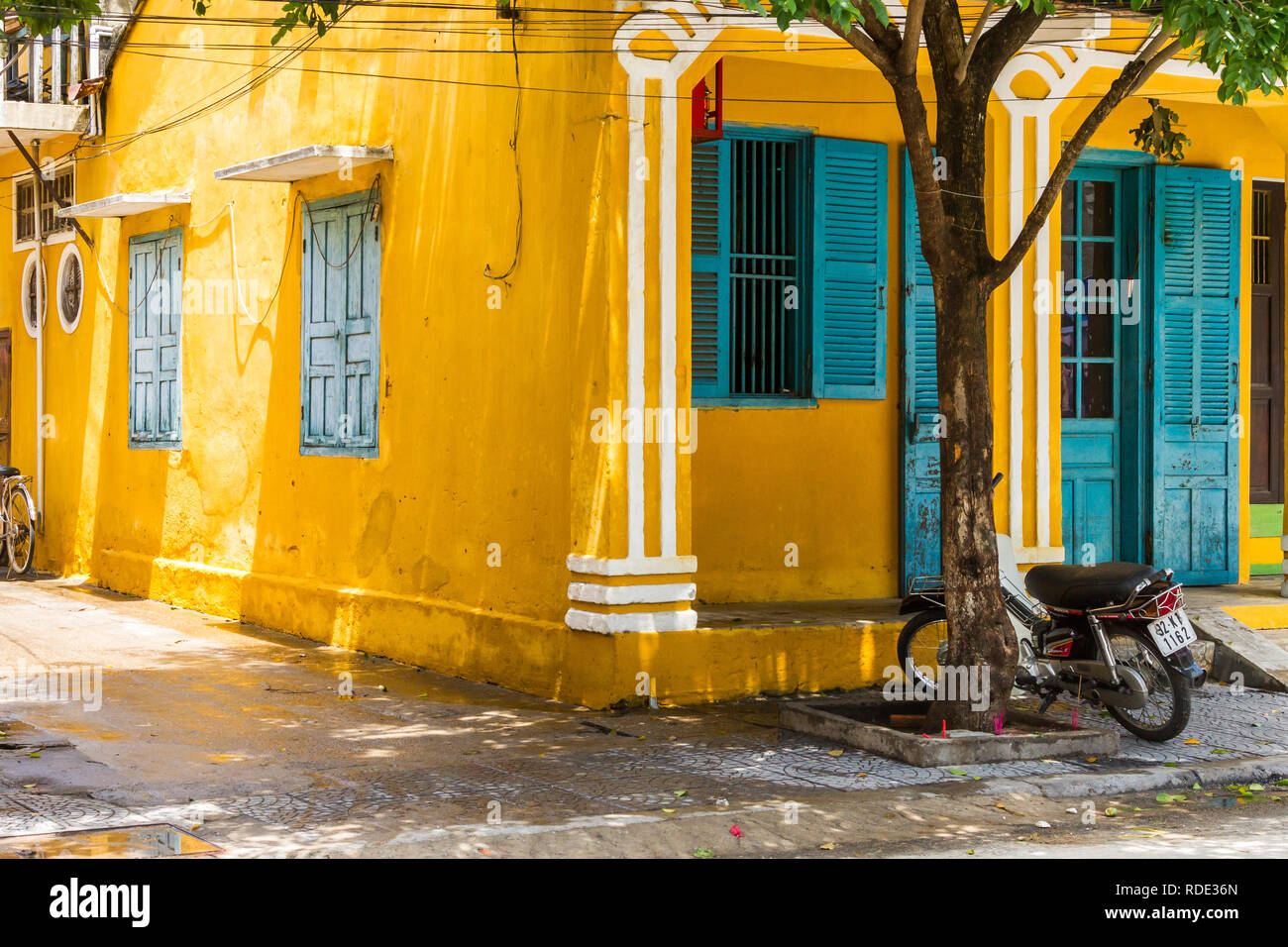 Vorder- und Seitenansicht leuchtend gelben Gebäude mit blauen Fensterläden und Türen in Hoi An, Vietnam mit Motorrad neben Baum Stockfoto