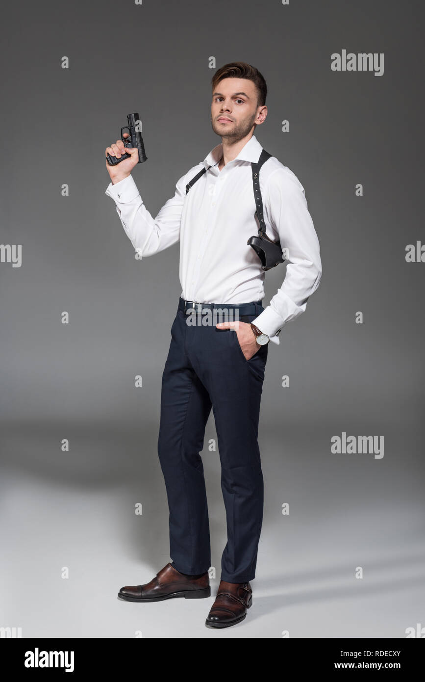 Hübscher junger Secret Agent mit Holster holding Pistole auf Grau Stockfoto