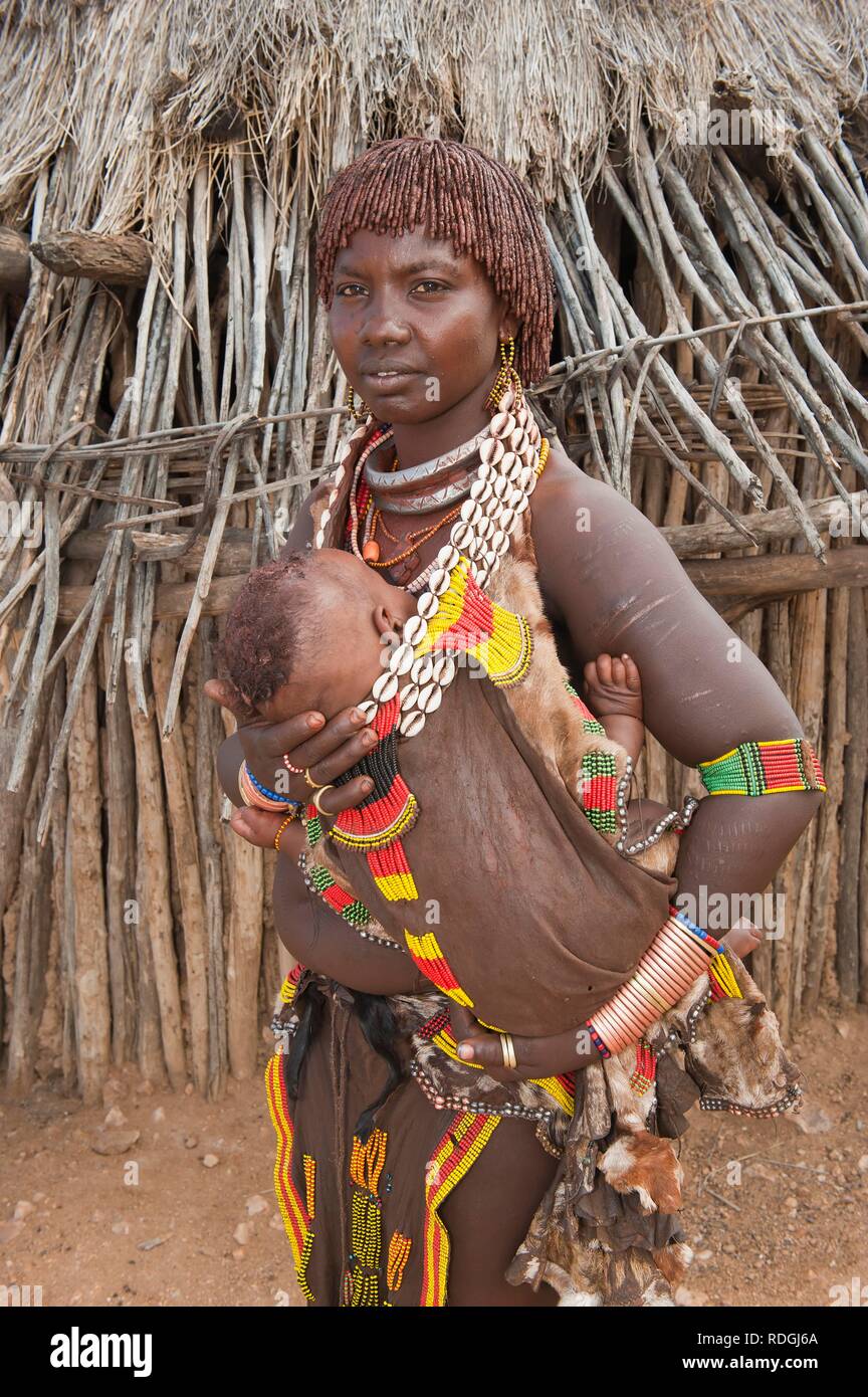 Junge Hamar Frau mit traditionellen Frisur mit rotem Lehm und Halsketten aus cowry Shells und ihr Baby in den Armen halten Stockfoto