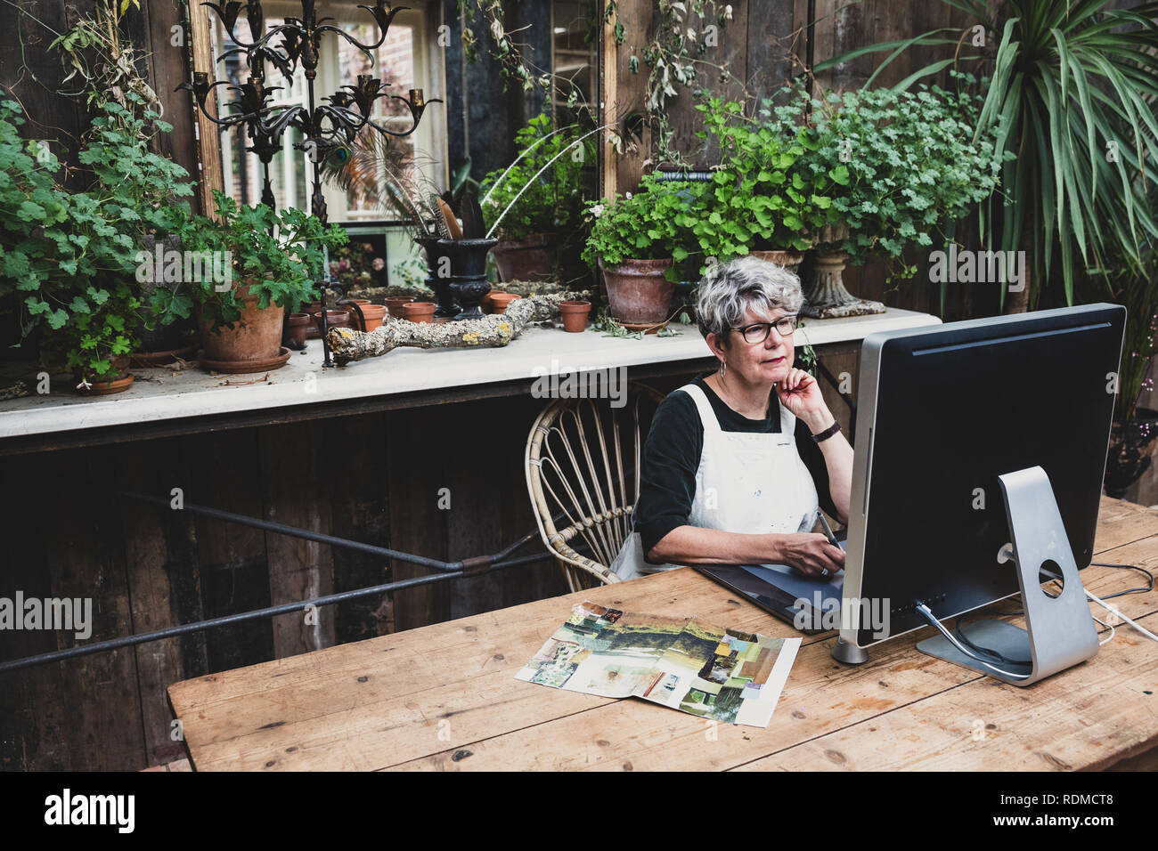 Ältere Frau Brille, schwarze und weisse Schürze an einem Holztisch sitzen, arbeiten auf dem Desktop Computer. Stockfoto