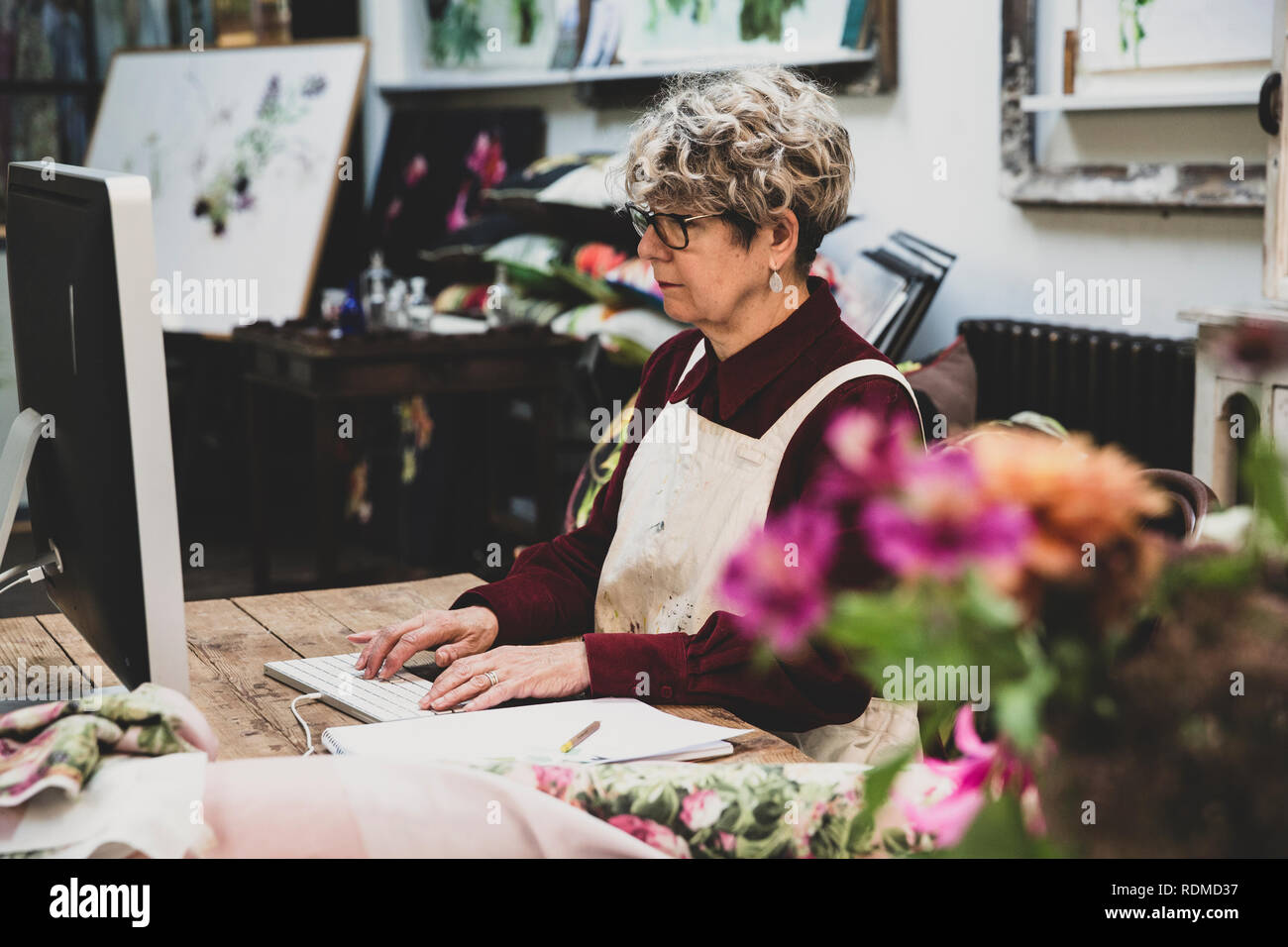 Ältere Frau Brille, rotes Kleid und weisse Schürze an den hölzernen Tisch sitzen in einem Studio, arbeiten auf dem Desktop Computer. Stockfoto