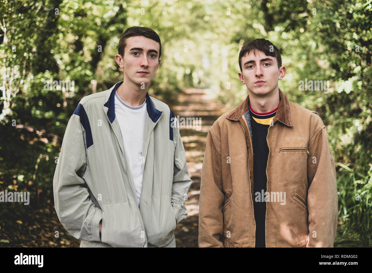 Junger Mann und Teenager mit kurzen braunen Haaren legere Jacken Seite an Seite auf einem Waldweg, an der Kamera schaut. Stockfoto