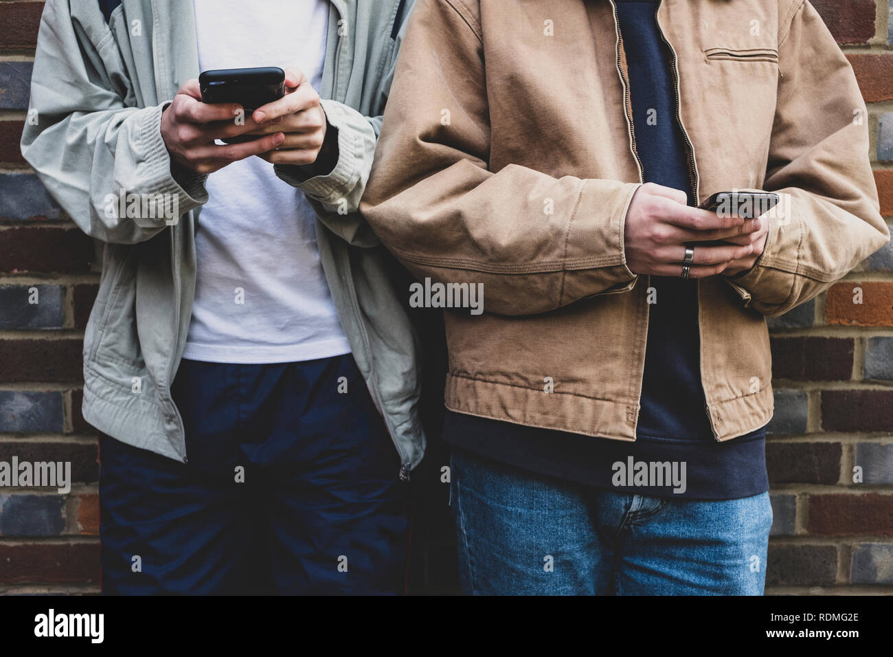 Den mittleren Abschnitt, zwei junge Männer legere Jacken Seite an Seite gegen die Wand, Textnachrichten auf Ihr Mobiltelefon. Stockfoto