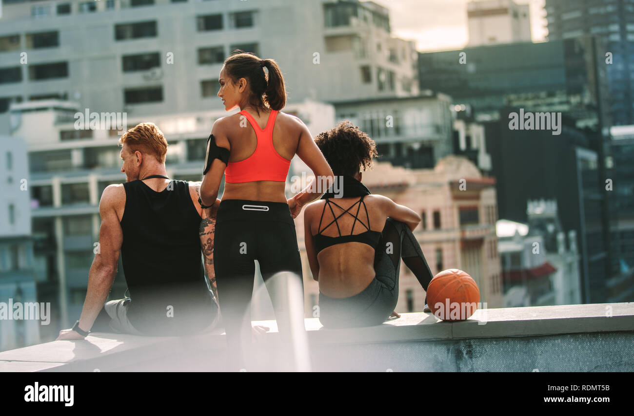 Fitnessmänner und -Frauen sitzen nach dem Fitnesstraining auf einem Dachzaun. Fitness-Leute, die sich nach dem Training entspannen, sitzen zusammen und schauen weg Stockfoto