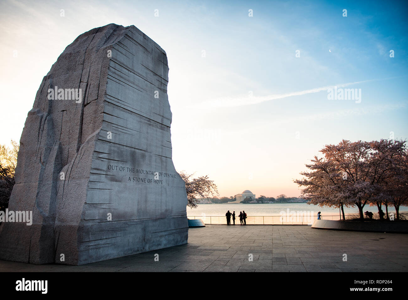 WASHINGTON DC – Kirschblüten blühen bei Sonnenaufgang in der Nähe des Martin Luther King Jr. Gedenkstätte am Tidal Basin. Die Granitskulptur „Stone of Hope“ des Gedenkwerks mit der Inschrift „Out of the Mountain of Desair, a Stone of Hope“ ist mit dem Jefferson Memorial in der Ferne über dem Wasser sichtbar. Stockfoto