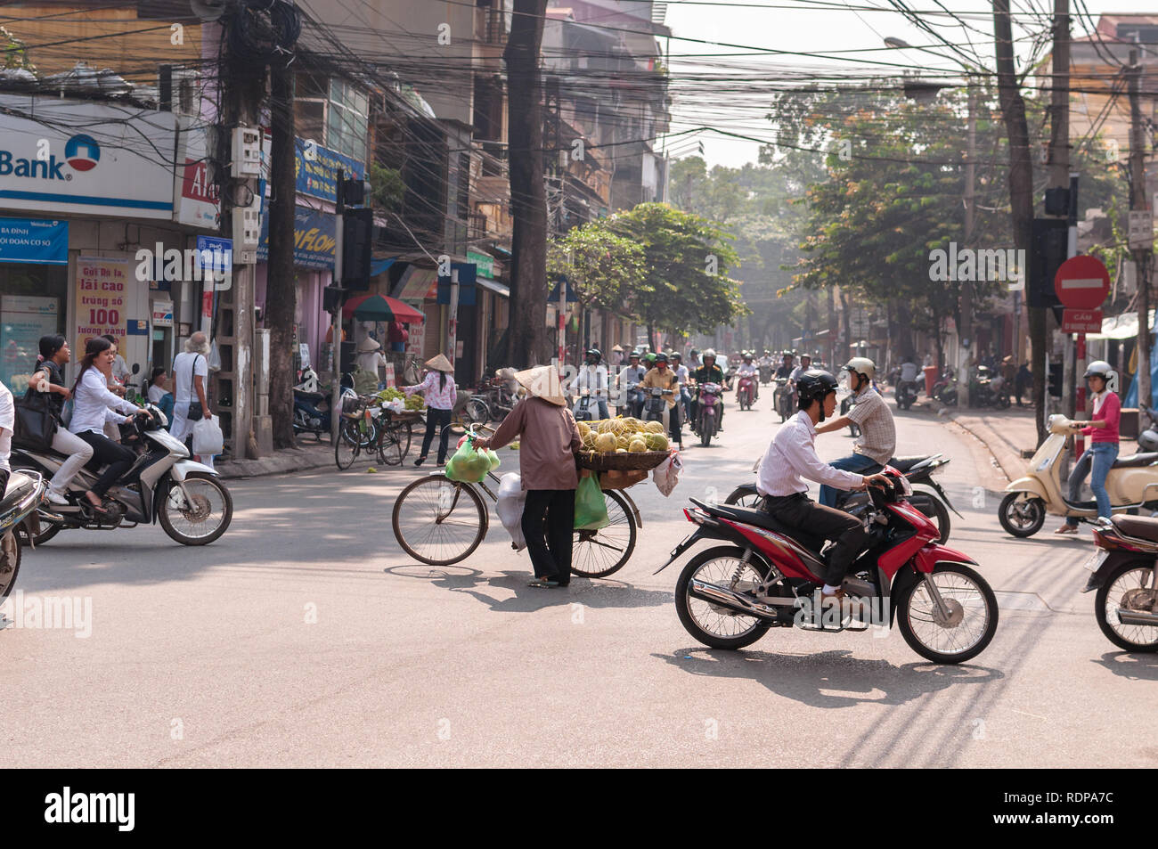 Eine vietnamesische Frau mit Fahrrad tragen Früchte warten eine Straße mit mehreren Mopeds und Motorräder, Hanoi, Vietnam zu überqueren Stockfoto