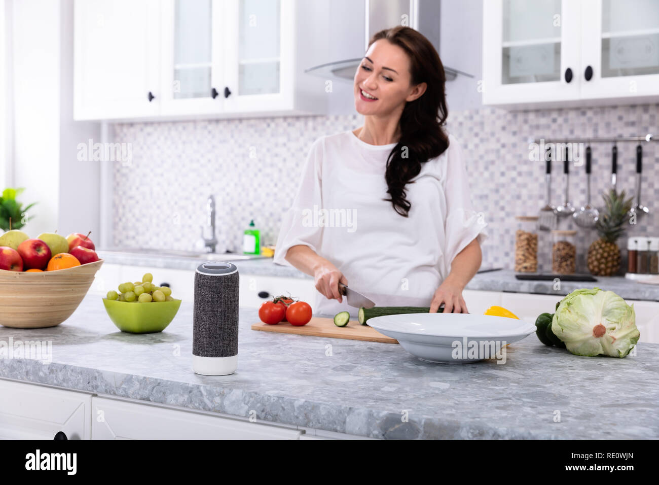 Glückliche Frau im Gespräch mit Voice Assistant beim Schneiden Gurken mit Messer Stockfoto