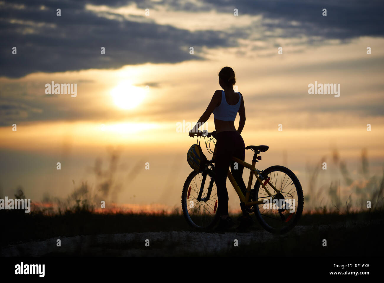 Rückansicht der Frau mit dem Fahrrad auf der Straße unter Gras genießen den Sonnenuntergang am Abendhimmel. Das Mädchen hat einen schönen sportlichen Körper. Helm hängt an den Griff des Fahrrad Stockfoto
