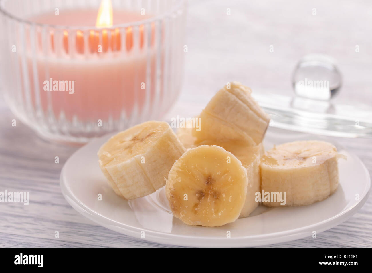 Bananenscheiben auf einem weißen Teller und einem leichten hölzernen Tisch, bereit für den Einsatz in der Ernährung. Rosa Kerze im Glas neben Schönheit und Frühstück lo Stockfoto