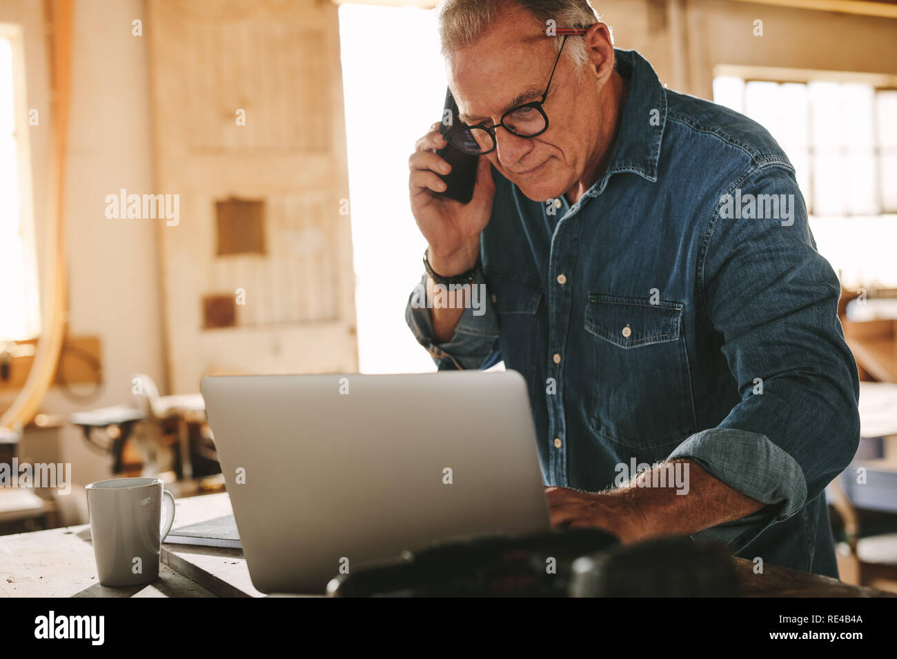 Reifen Mann, besetzt mit Laptop und Gespräch am Handy in seiner Werkstatt. Ältere männliche Tischler arbeiten, die auf Notebook- und Anruf in seinem carpen Stockfoto