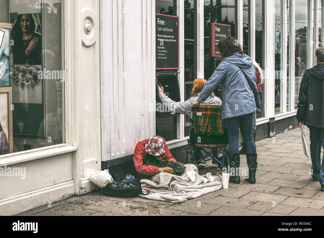 Ein Mann ist betteln auf der Straße, während eine Frau in einem Rollstuhl ist für Bargeldbezug an der High Street in Großbritannien Stockfoto