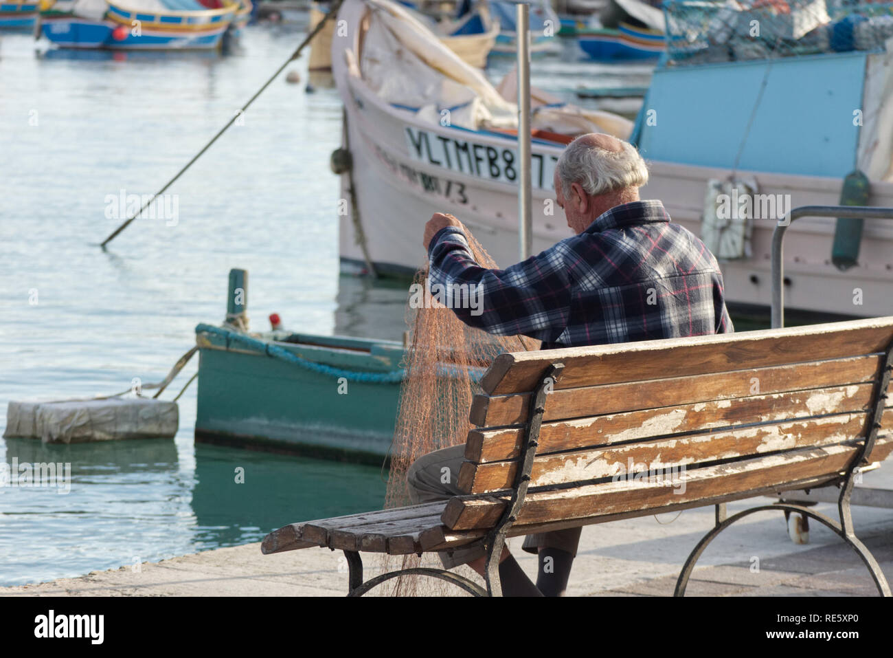 Marsaxlokk/Malta - 30. November 2018: Ein alter Fischer ist die Reparatur seiner Fischernetze auf einer Bank am Hafen. Stockfoto