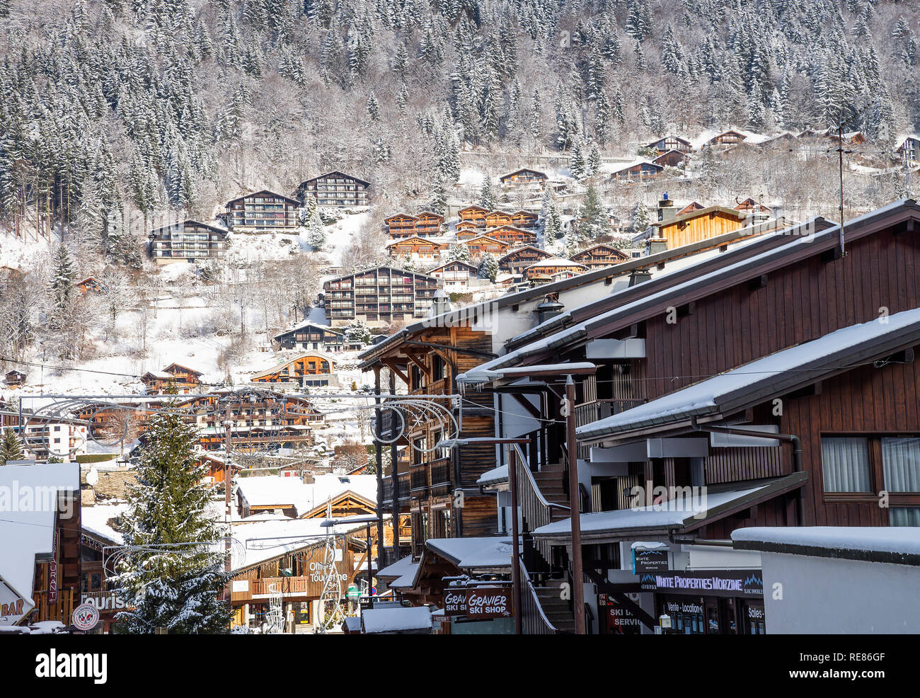 Geschäfte und Restaurants mit bunten Zeichen mit Chalets am Hang und Verschneiten Kiefernwald in Morzine Haute Savoie Portes du Soleil Frankreich Stockfoto