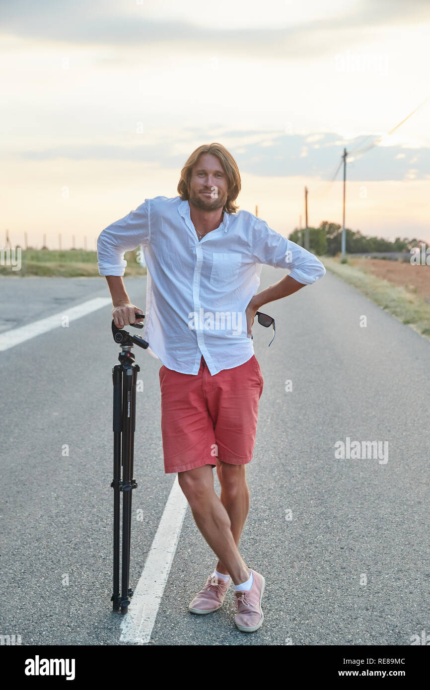 Die schöne junge brutalen Mann steht auf der Straße bei Sonnenuntergang, er ist in ein weißes Hemd mit kurzen Ärmeln und rote Shorts gekleidet, der Fotograf Stockfoto