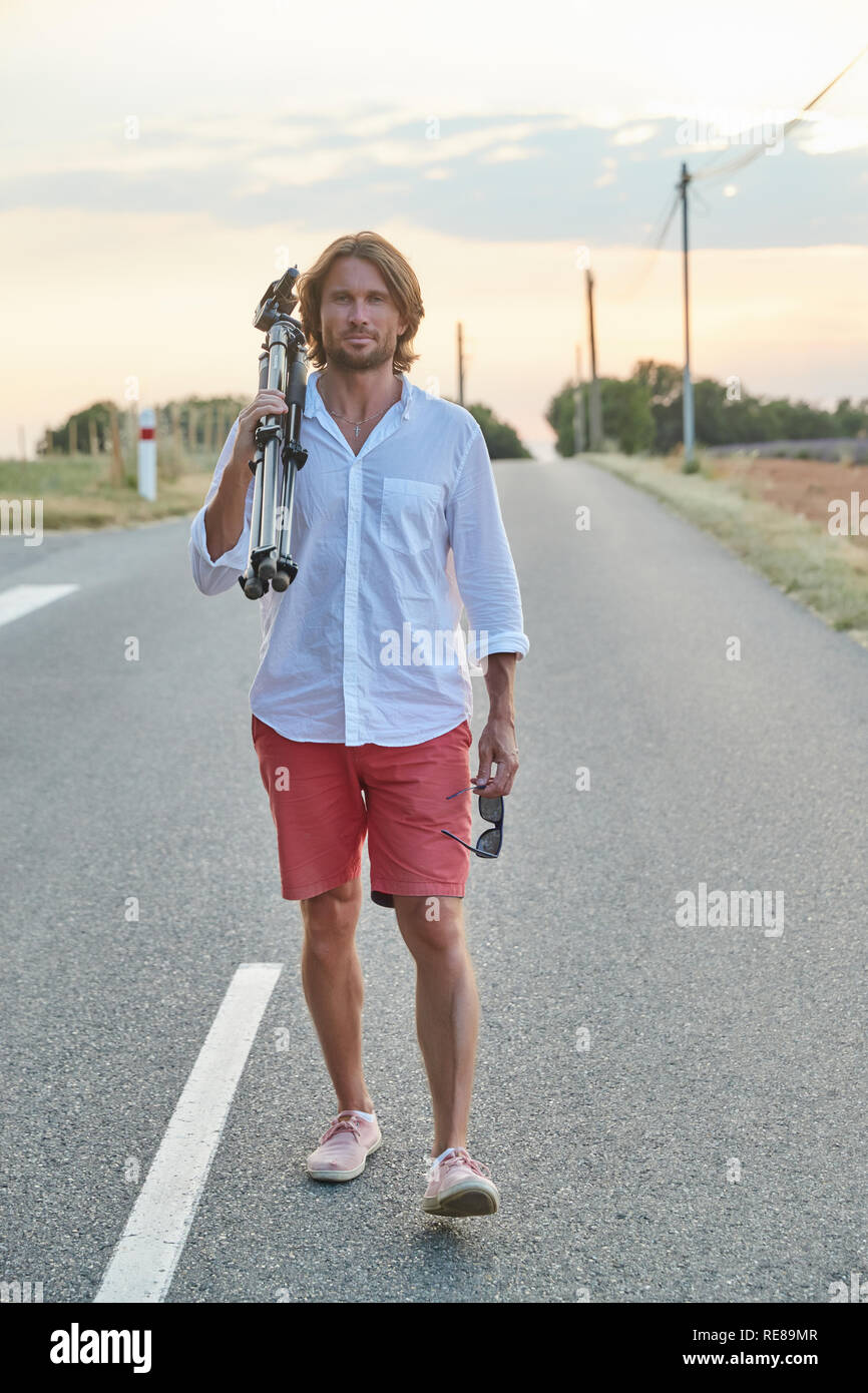 Die schöne junge brutalen Mann steht auf der Straße bei Sonnenuntergang, er ist in ein weißes Hemd mit kurzen Ärmeln und rote Shorts gekleidet, der Fotograf Stockfoto