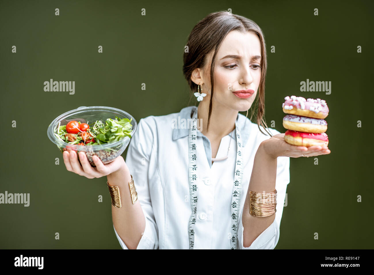 Junge Frau Ernährungsberaterin auf die Krapfen mit traurigen Emotionen Wahl zwischen Salat und ungesunden Nachtisch auf dem grünen Hintergrund Stockfoto
