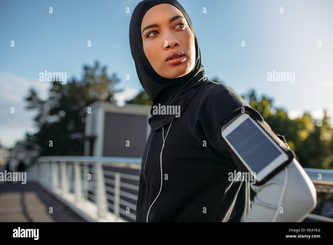 Junge Sportlerin im hijab stehen im Freien mit Mobiltelefon auf Ihr Armband weg schauen. Gesunde weibliche eine Pause nach dem Training im Freien in Stockfoto