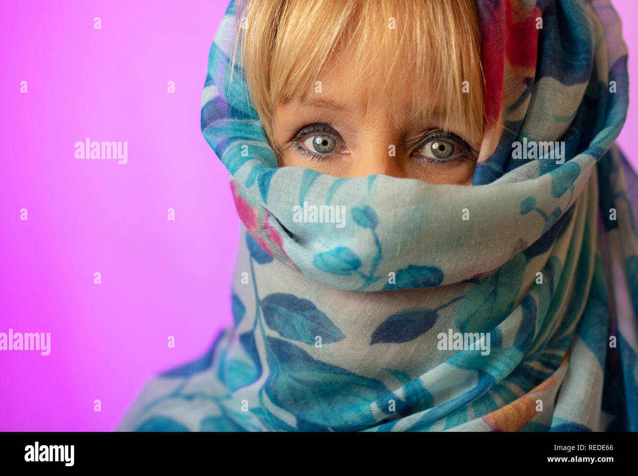 Weiße Frau mit blonden Haaren und blauen Augen trägt einen blauen Kopftuch  als modisches Accessoire mit nur Augen und Fringe zeigt vor einem rosa  Hintergrund Stockfotografie - Alamy