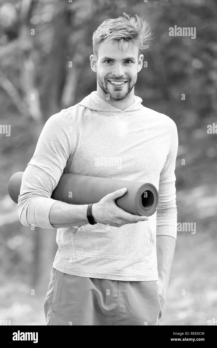 Sportler Training mit Yoga Matte. Zeit für Yoga Konzept. Mann Athlet auf glückliches Gesicht trägt Yoga Matte, Stretching, Natur Hintergrund. Sportler mit Fitness tracker und Matte für Stretching. Stockfoto