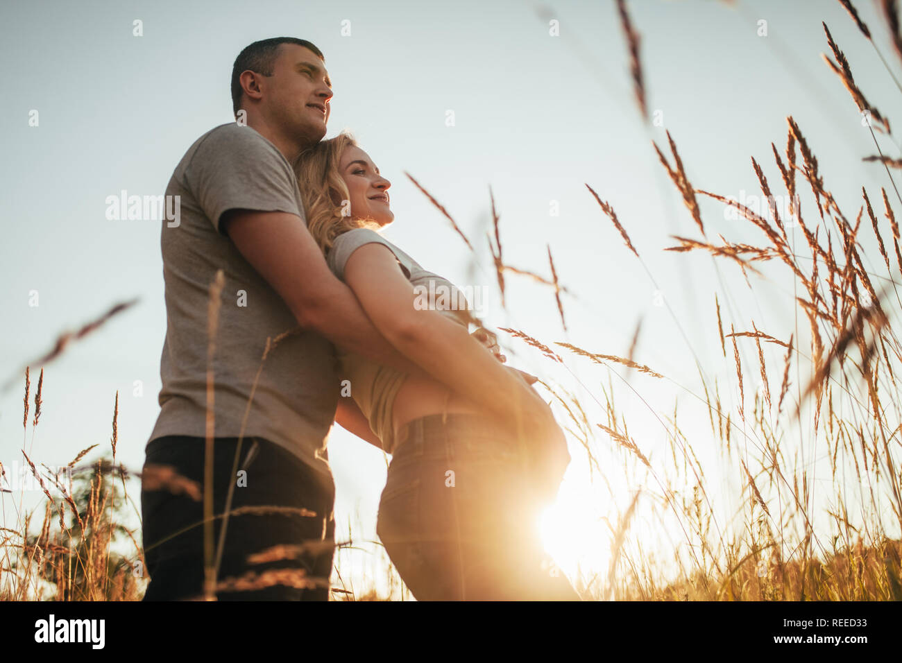 Ein junger Mann umarmt den Bauch der seine schwangere Frau bei einem Spaziergang gegen den Hintergrund von Himmel und ährchen. Hintergrundbeleuchtung. Stockfoto