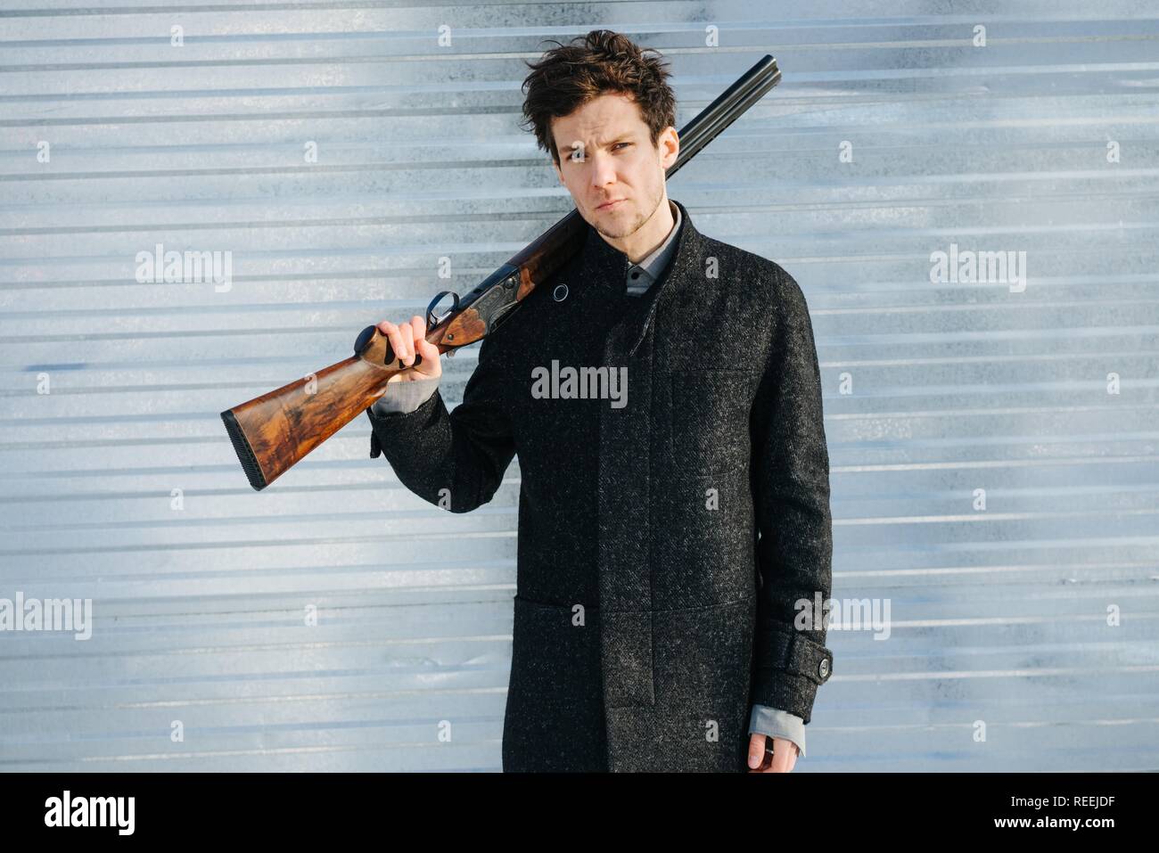 Porträt einer stilvollen ernster Mann in Mantel mit Doppelflinte auf der Schulter an einem sonnigen Wintertag. Blech zaun Hintergrund. Blick auf die Stockfoto