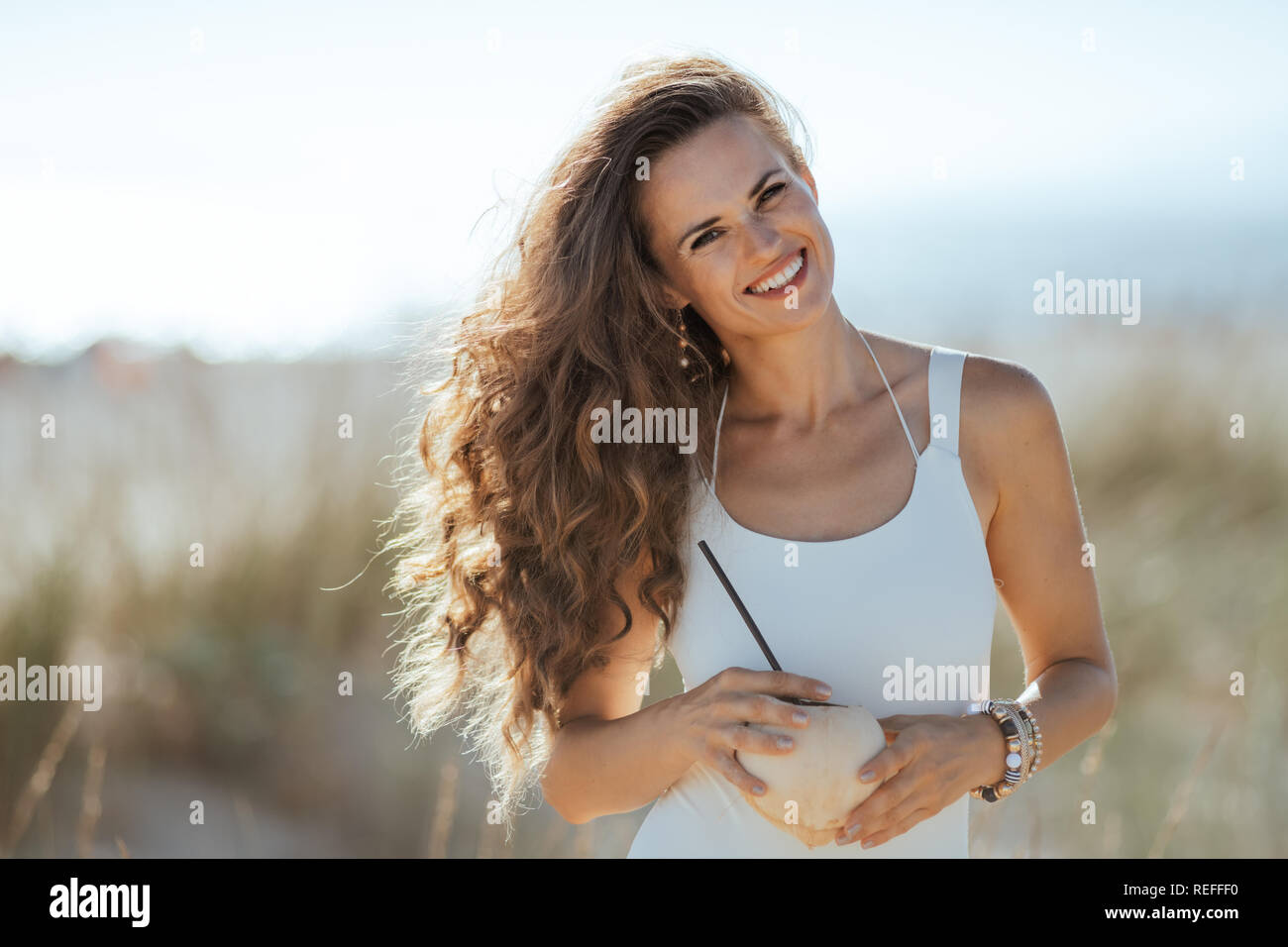 Portrait von lächelnden jungen Frau in Weiß beachwear Holding Kokosnuss mit Stroh auf den Strand. Nutzen für die Gesundheit der Kokosnuss Früchte sind zahlreich. minimal Stockfoto