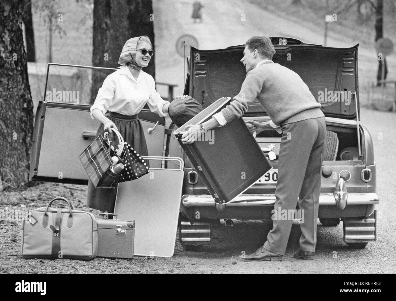 Paar 1958. Ein junges Paar packt den Kofferraum seines Autos mit Gepäck und Campingausrüstung für einen Autourlaub. Ein praktischer Klapptisch ist ebenfalls verpackt. Sie trägt ein typisches Herrenhaus aus den 1950er Jahren mit einem weißen Hemd, einem breiten Rock, einer Sonnenbrille und einem Schal. Schweden 1958 Stockfoto