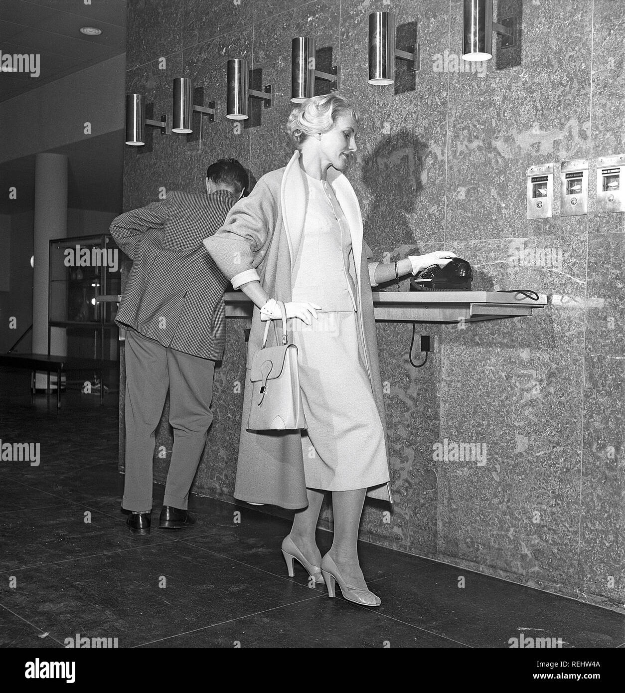 Frau in den 1950er Jahren. Ein junges Paar in einem Flughafen, ist über das Telefon. Sie ist modisch in einem typischen 50er Jahre zwei Stück kleid mit passendem Rock und Jacke gekleidet, mit einem leichten Mantel. Die Handschuhe, Schuhe und Tasche sind in Übereinstimmung zu bringen. Schweden 1958. Foto Kristoffersson Ref CB 15-7 Stockfoto