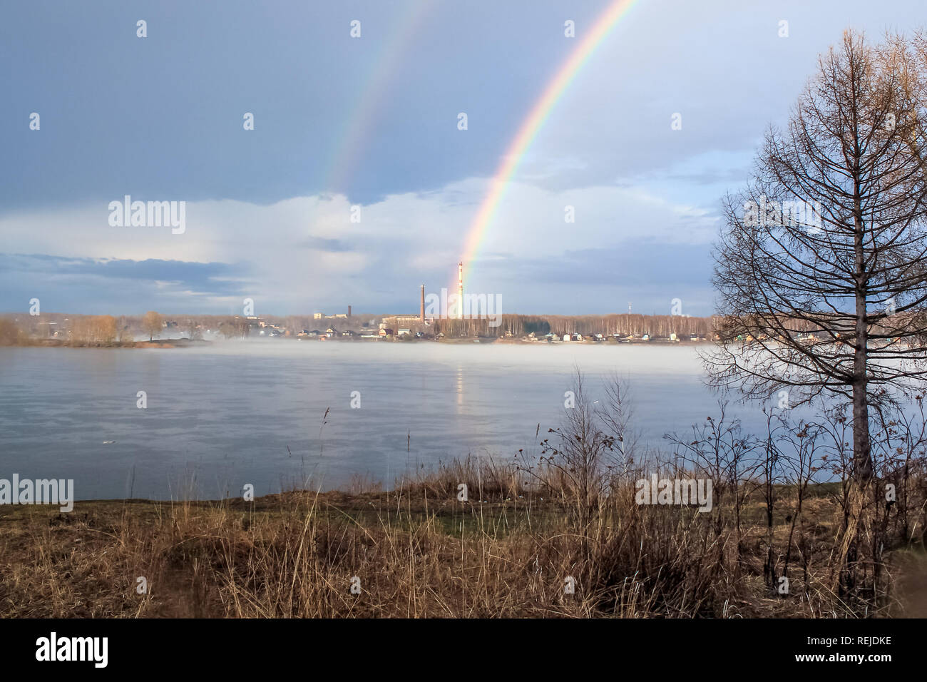 Friedlichen Frühling neblige Landschaft bei Sonnenaufgang nach dem Regen mit Regenbogen auf dem Fluss, im Hintergrund Industrieanlagen mit Rohr Stockfoto