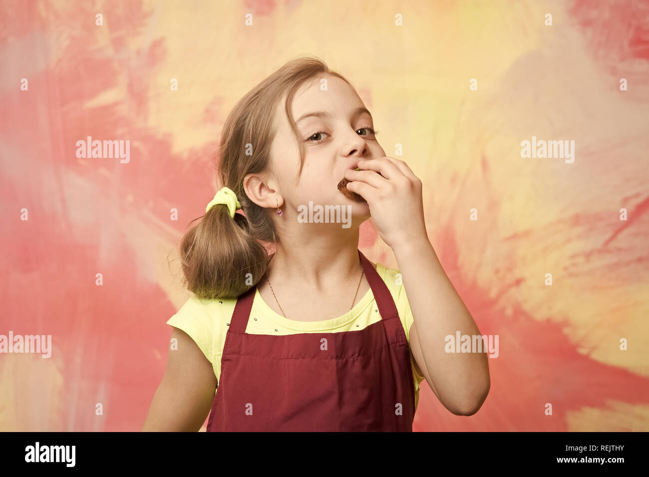 Kind beißen Kuchen. Mädchen essen Kuchen. Gesunde Ernährung und Diät Konzept. Backen und köstliche Backwaren. Baby Cook in rot Koch Schürze auf farbenfrohe abstrakte Wand. Stockfoto