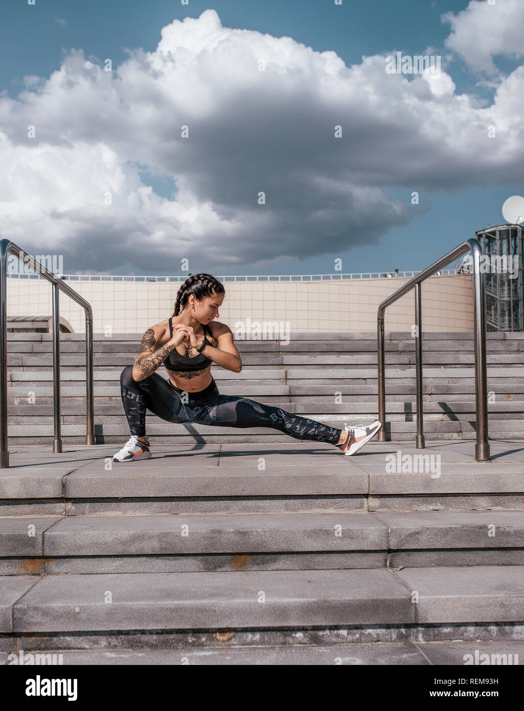 Mädchen Athlet ist Stretching, vor dem Joggen am Morgen in der Stadt auf den Stufen. Vor dem Hintergrund der konkreten und Wolken. Gesunde Lebensweise. In Sportkleidung und Turnschuhe. Gebräunte Haut und Tätowierungen. Stockfoto