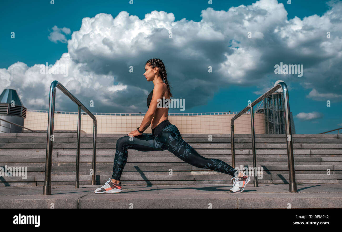 Athlet Mädchen tun Stretching vor dem Joggen am Morgen in der Stadt. Vor dem Hintergrund der konkreten Treppen und Wolken. Das Konzept einer gesunden Lebensweise. In Sportkleidung und Turnschuhe. Stockfoto