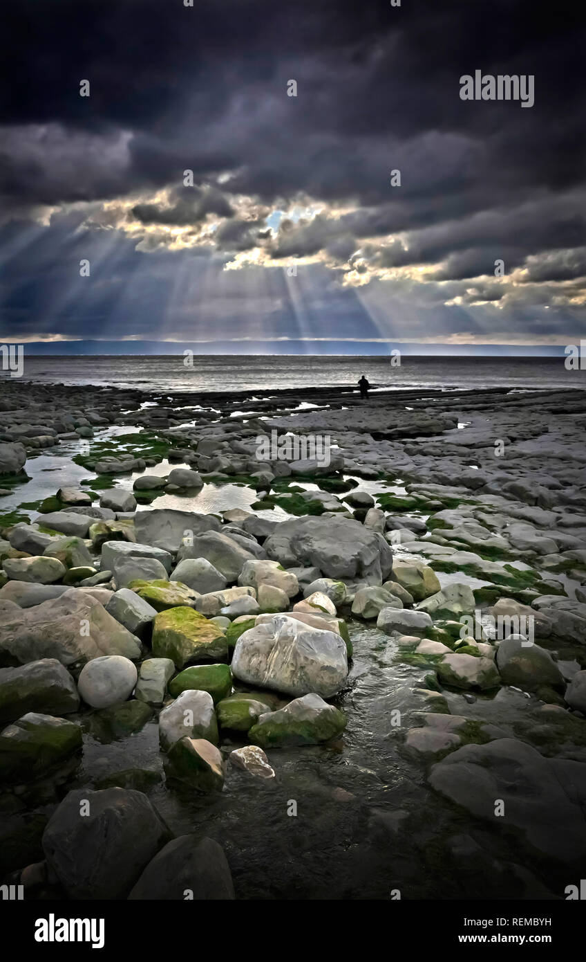 Felsiger Strand mit stürmischem Himmel und dramatischem Licht und eine Person in Silhouette in der Ferne durch Wasser Rand Einsamkeit Konzept Stockfoto
