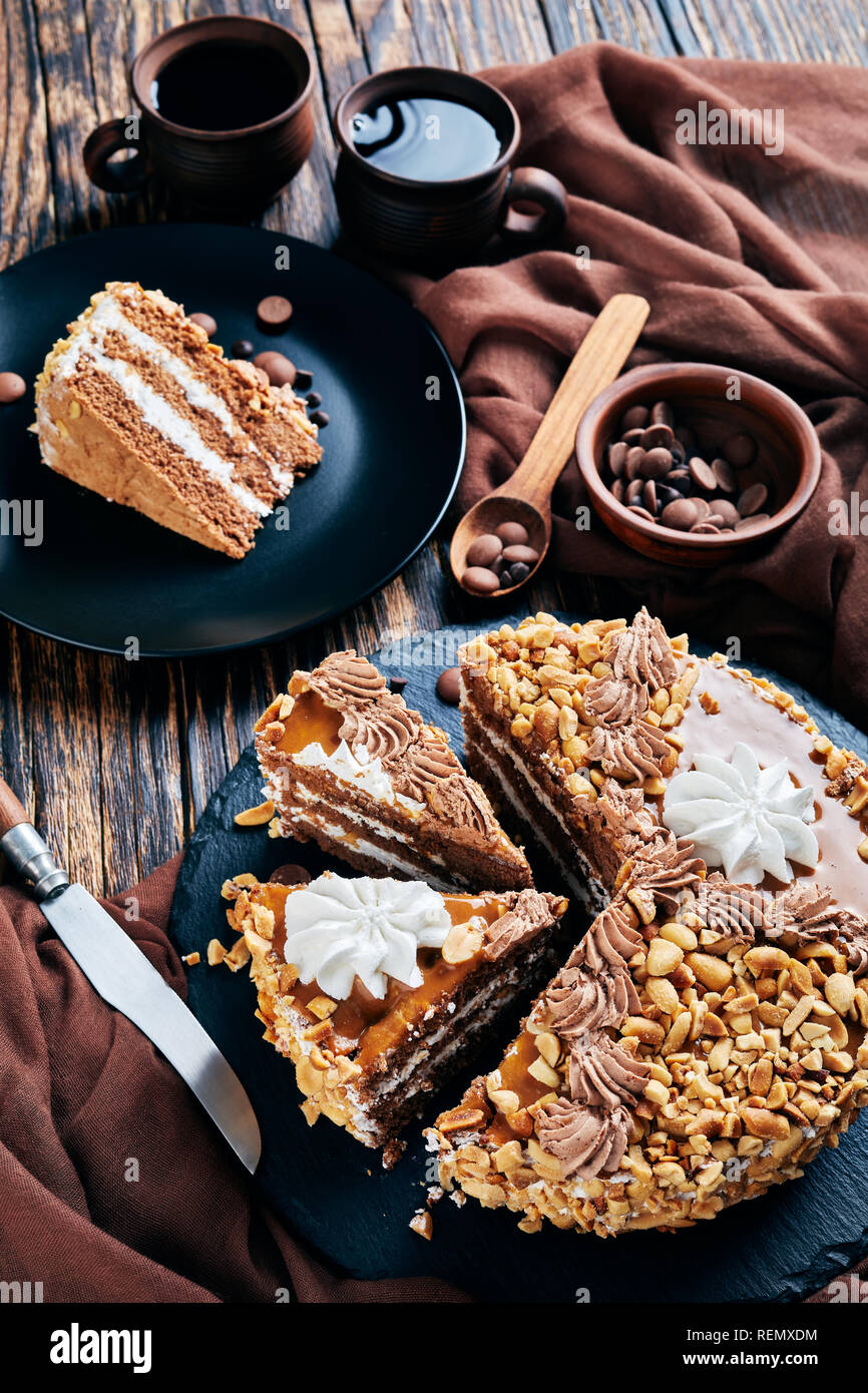 Erdnuss Kuchen gekrönt mit Kakao Peanut Butter, Sahne und Eischnee Rosen auf schwarzem Teller auf einem rustikalen Holztisch mit Kaffee aus Steingut Schale serviert. Stockfoto