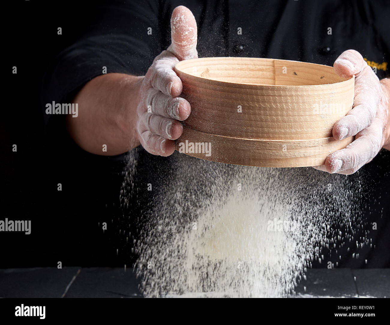 Man siebt weißes Mehl durch ein Sieb, schwarzer Hintergrund Stockfoto