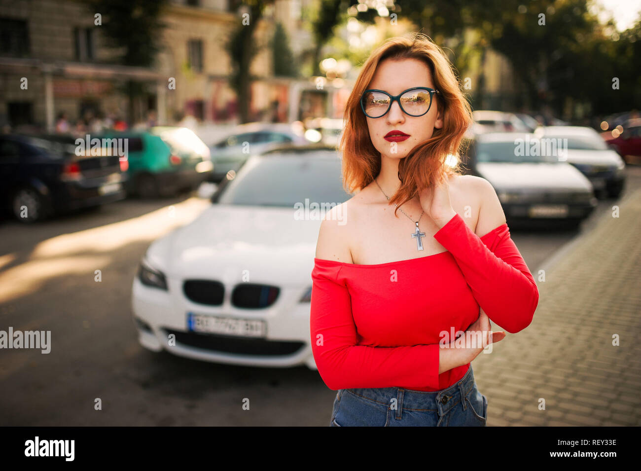 Attraktive rothaarige Frau in Brillen, Verschleiß auf rote Bluse und Jeans Rock auf der Straße gegen weiße Sport Auto posiert. Stockfoto