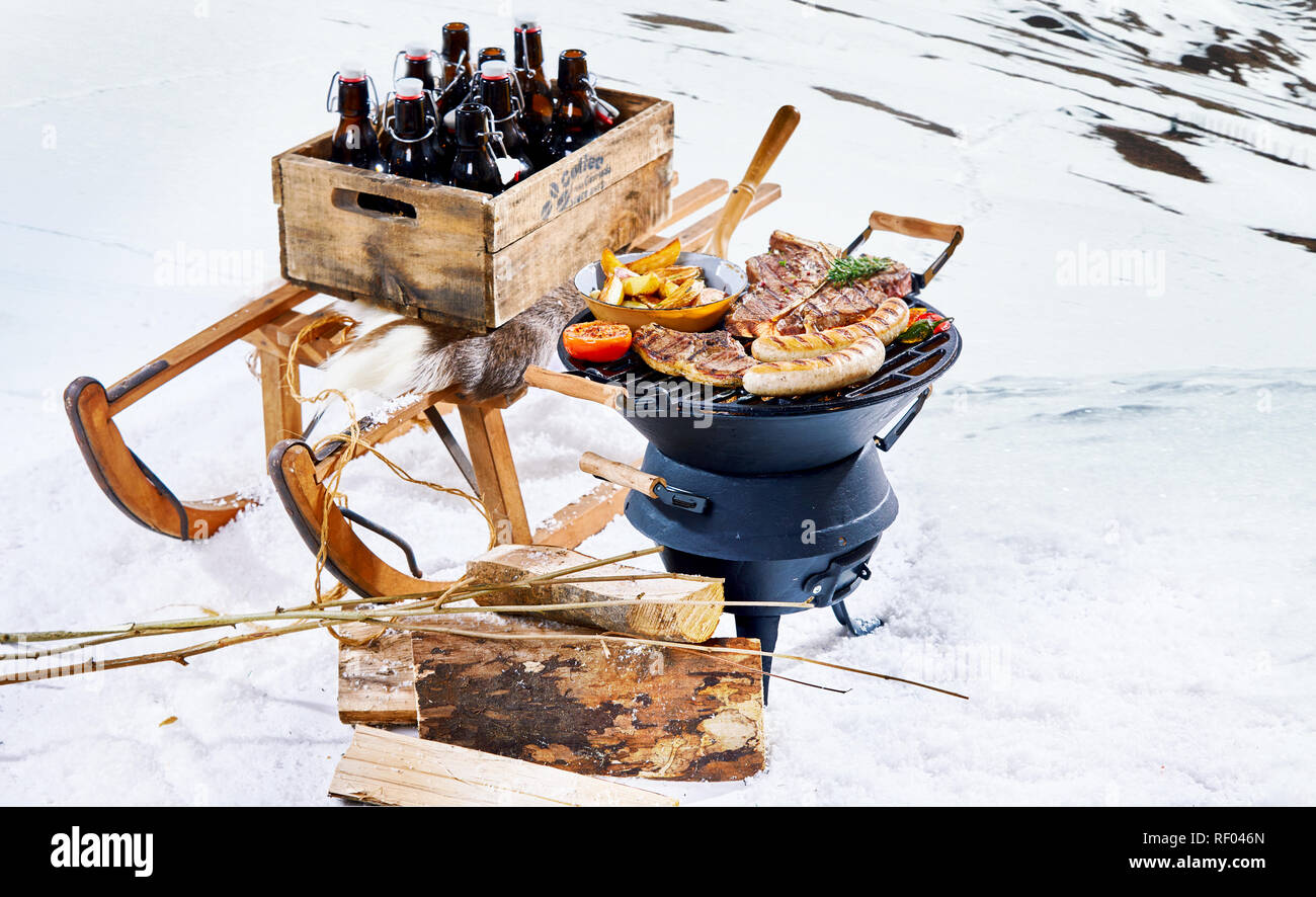 Schneereiche Winter Grill draußen in der Kälte mit einer Kiste Bier  Flaschen auf einem Schlitten und würzigen Würstchen und T-Bone Steaks  grillen auf einem tragbaren Grill Stockfotografie - Alamy