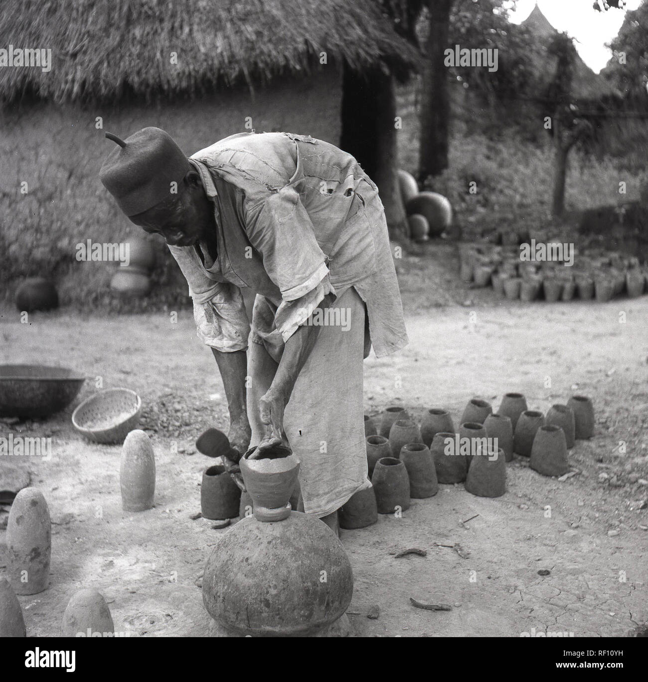 1950 s, historischen, lokalen afrikanischen tragen traditionelle Yoruba Hut, Aso Oke Hut und Tuch Robe, die Tongefäße, die außerhalb seiner Hütte, Nigeria, Afrika. Stockfoto