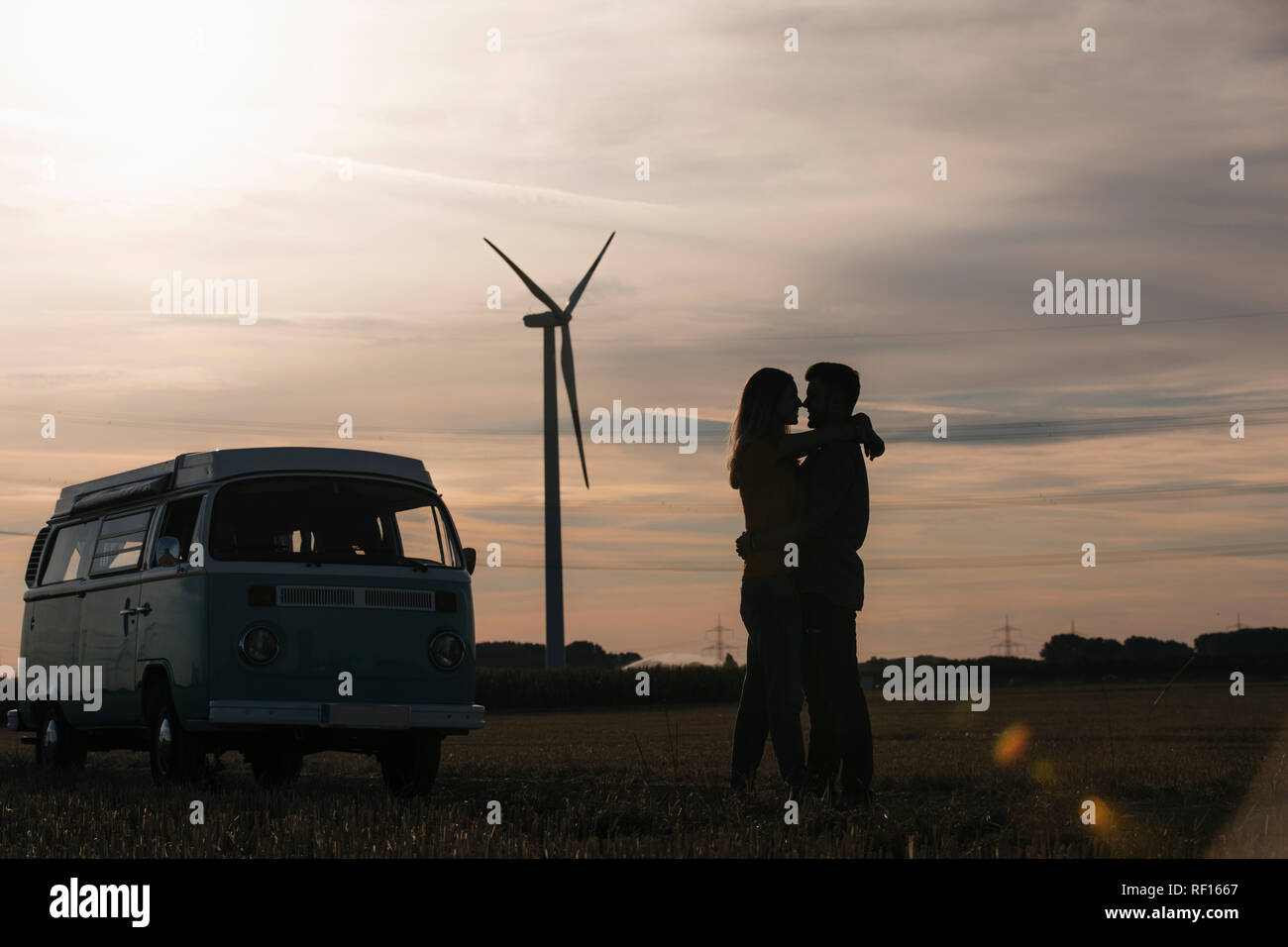 Junges Paar Küssen an Camper van in der Dämmerung mit Windenergieanlage im Hintergrund Stockfoto