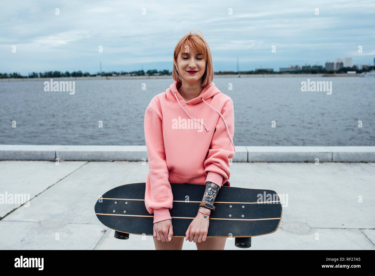 Lächelnde junge Frau mit Carver skateboard Stehend am Flußufer Stockfoto