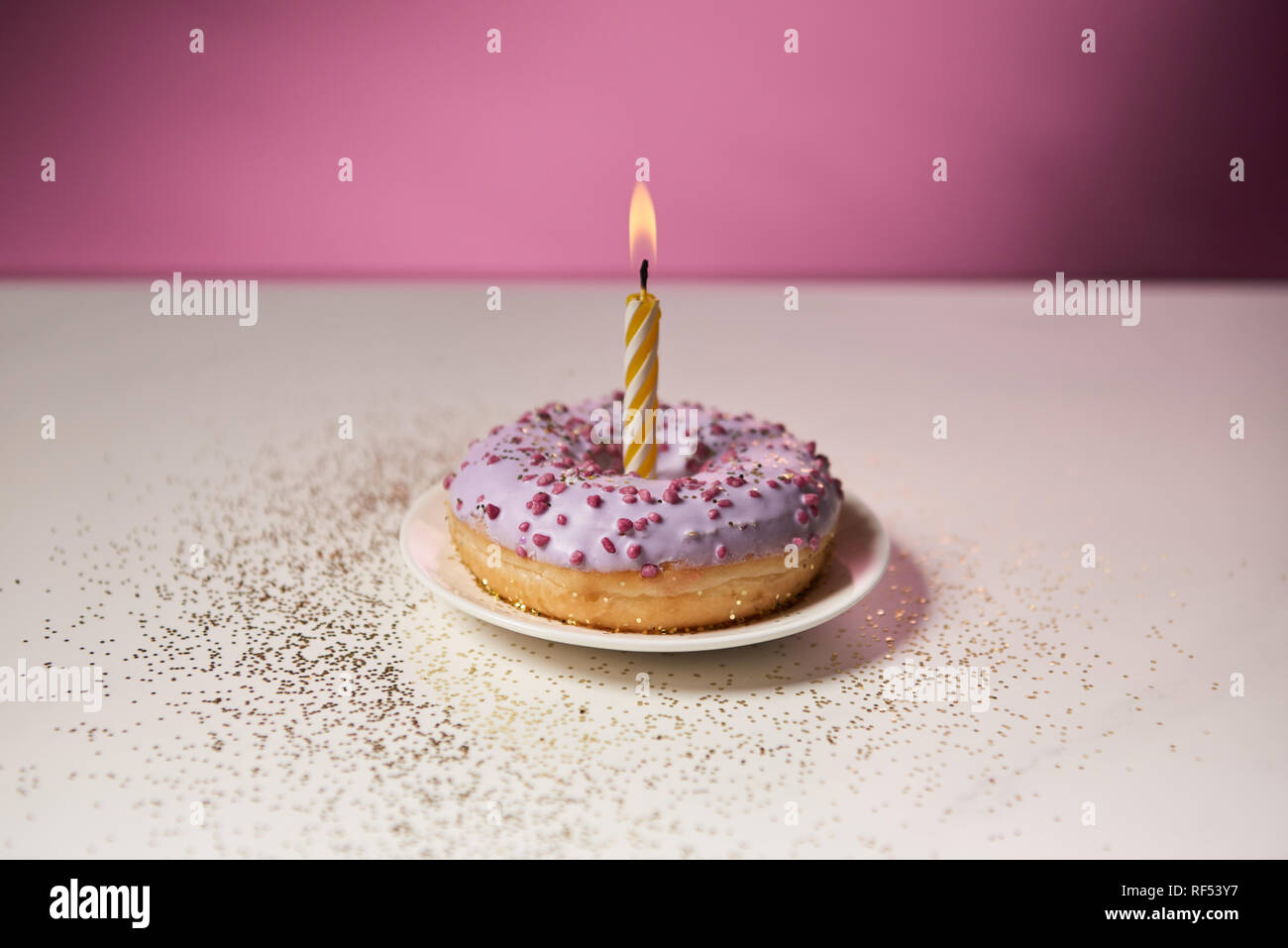 Brennende Kerze in der Mitte der glasierten Krapfen mit funkelt auf weißen Tisch auf rosa Hintergrund Stockfoto