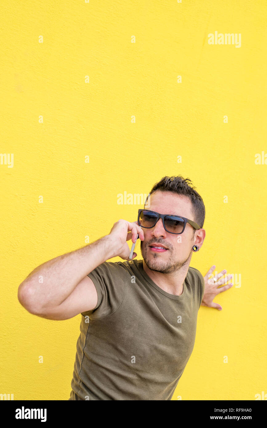 Vorderansicht eines jungen erwachsenen Mann gegen eine gelbe Wand mit Sonnenbrille, während Sie ein Handy im Freien an einem sonnigen Tag Stockfoto